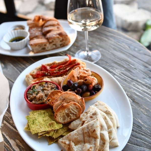 Appetizer platter with various dips, olives, pita bread, and baguette slices, with a glass of white wine.