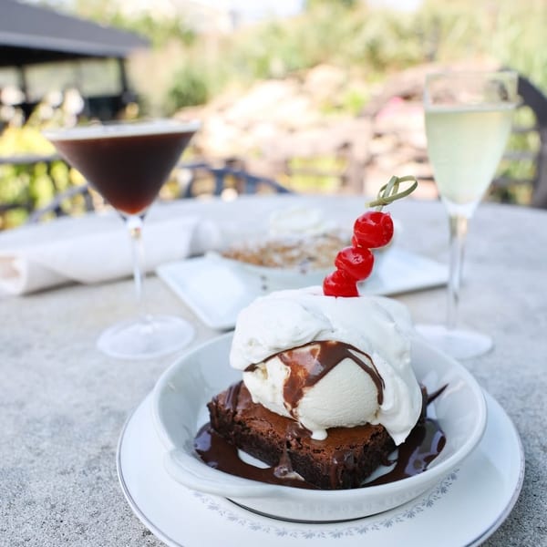 Brownie with vanilla ice cream, chocolate sauce, and cherries, with drinks on an outdoor table.