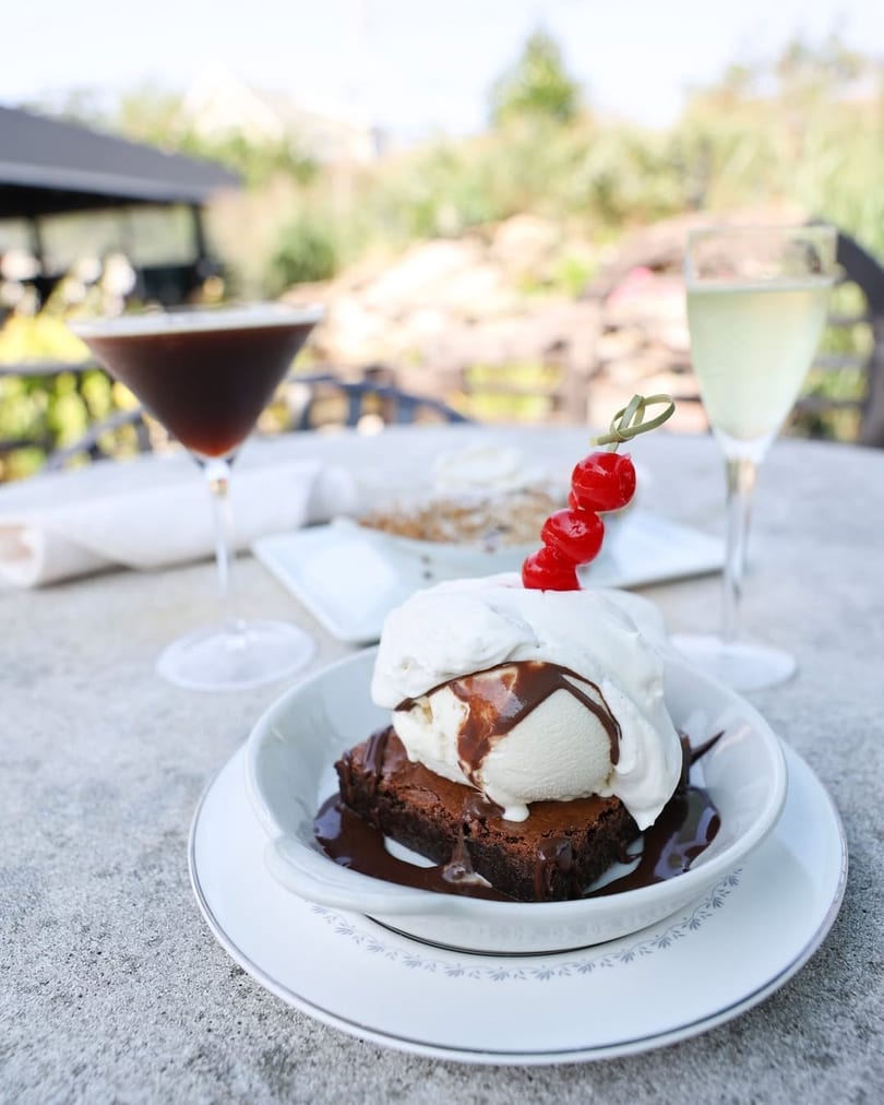 Brownie with vanilla ice cream, chocolate sauce, and cherries, with drinks on an outdoor table.