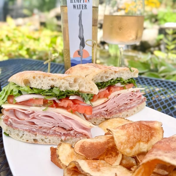 Deli sandwich with chips, a bottle of Hampton Water rosé, and a glass of wine outdoors.